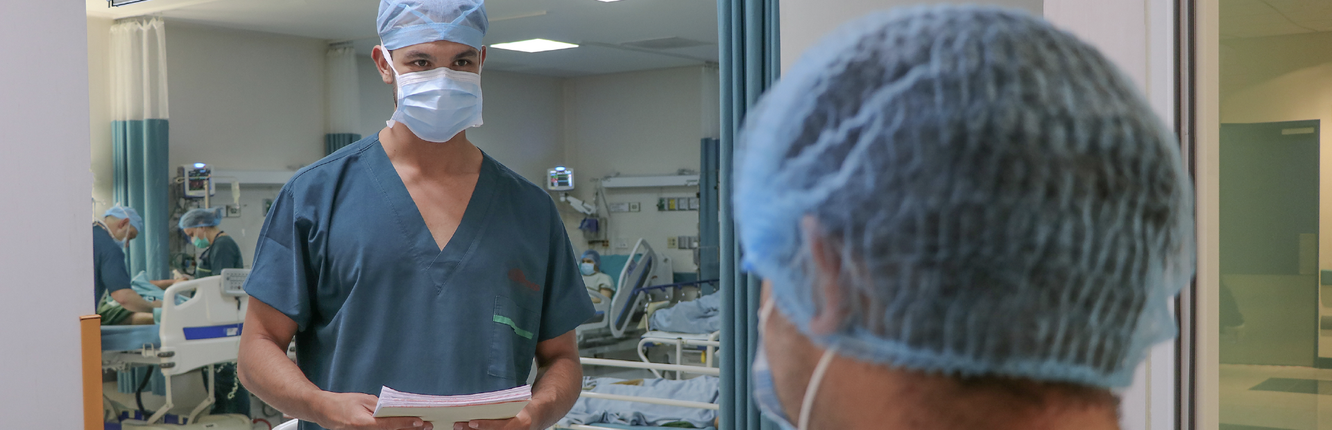 En primer plano se ve un paciente de pie de espalda y frente a él un enfermero recibiéndole en el pasillo quirúrgico
