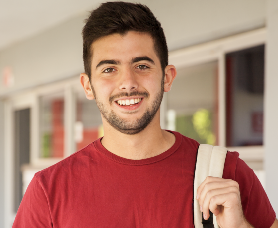 Joven de universidad, con bulto en un hombro, sonriendo a cámara