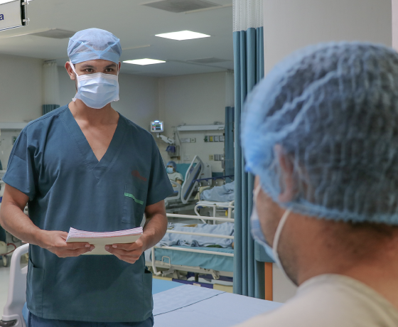 En primer plano se ve un paciente de pie de espalda y frente a él un enfermero recibiéndole en el pasillo quirúrgico