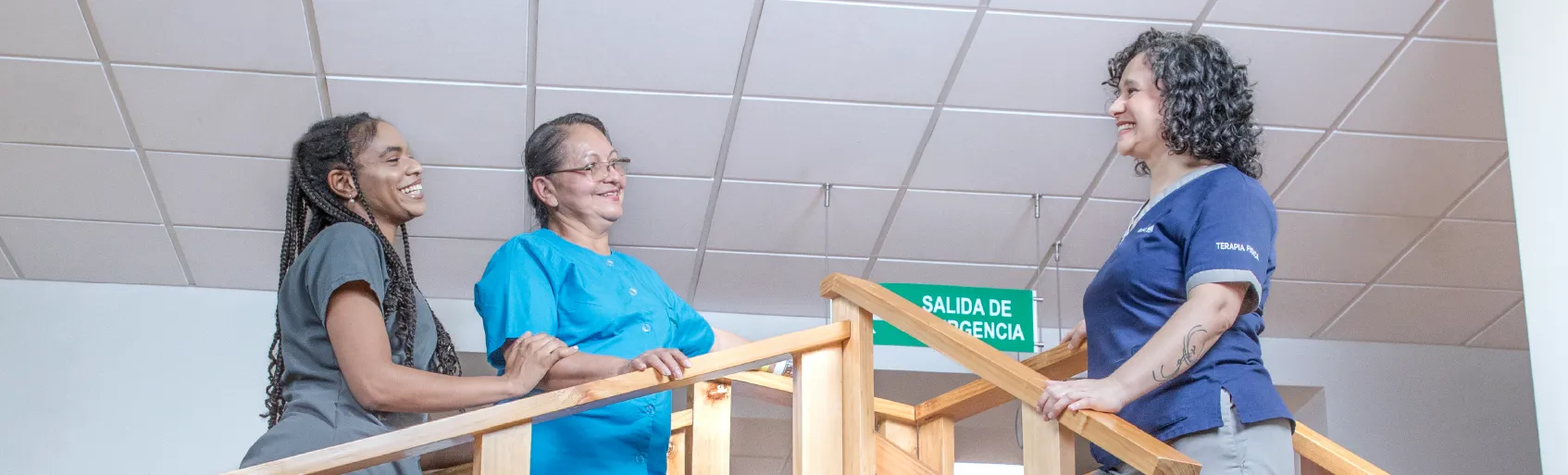 Fisioterapeuta sonríe a paciente que es cuidada por asistente de pacientes, las tres personas se encuentran de pie sobre escaleras de terapia física