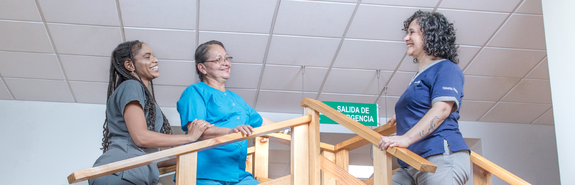 Fisioterapeuta sonríe a paciente que es cuidada por asistente de pacientes, las tres personas se encuentran de pie sobre escaleras de terapia física